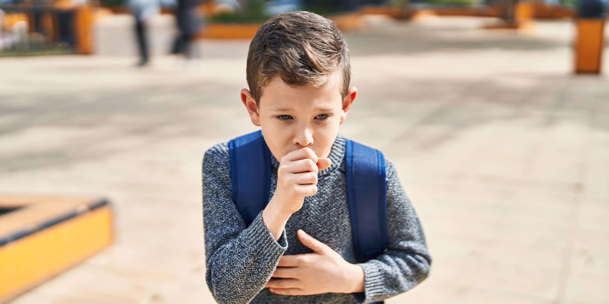 A child wearing a backpack coughs into his closed fist.