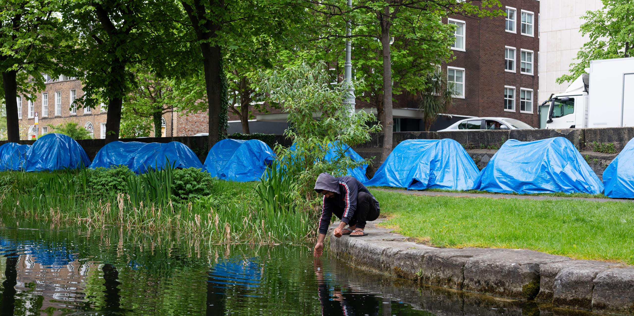 A man squats next to the Grand Canal in Dublin and washes his hands, while a row of blue tents is visible behind him on the canal bank
