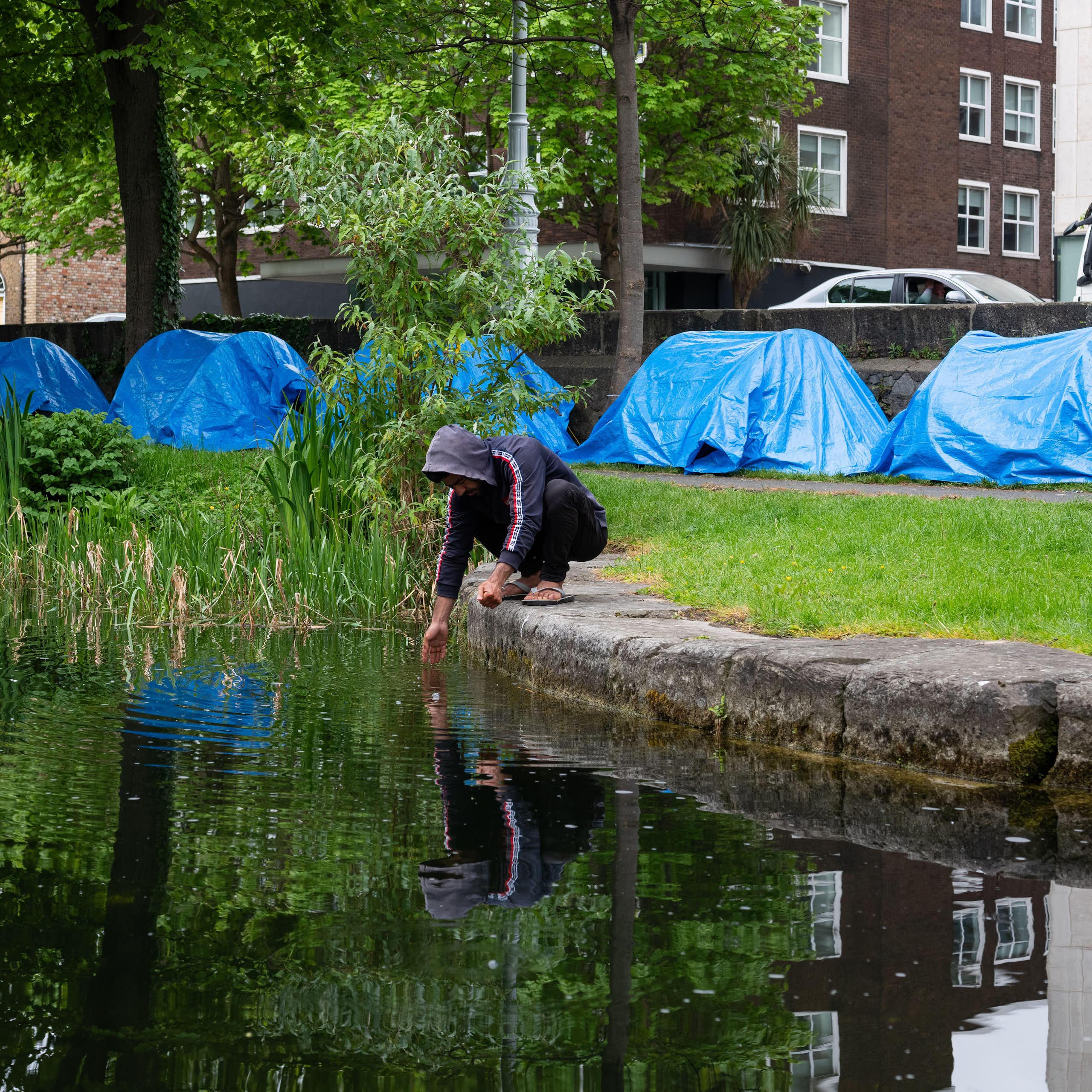 A man squats next to the Grand Canal in Dublin and washes his hands, while a row of blue tents is visible behind him on the canal bank