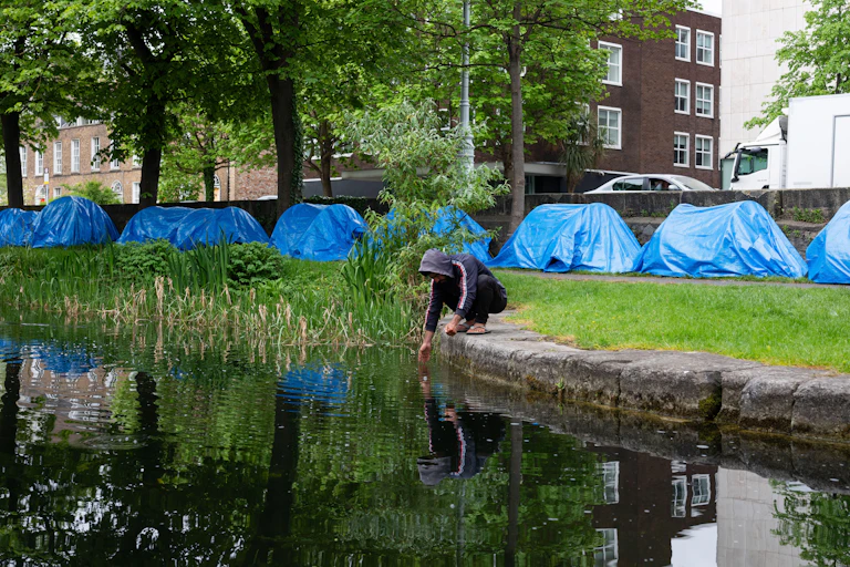 A man squats next to the Grand Canal in Dublin and washes his hands, while a row of blue tents is visible behind him on the canal bank