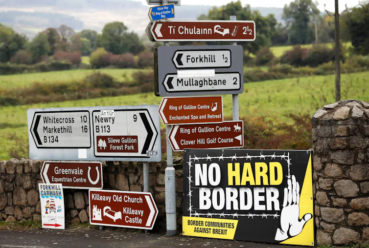 View of a collection of traffic signs against a stone wall, with one large sign that reads 'no hard border' and has an image of a hand and barbed wire fence.