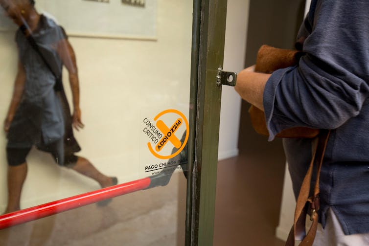 Uma porta de vidro com um adesivo amarelo onde se lê 'addiopizzo'