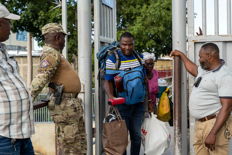 A man laden with bags walks through a metal door flanked by two security guards.