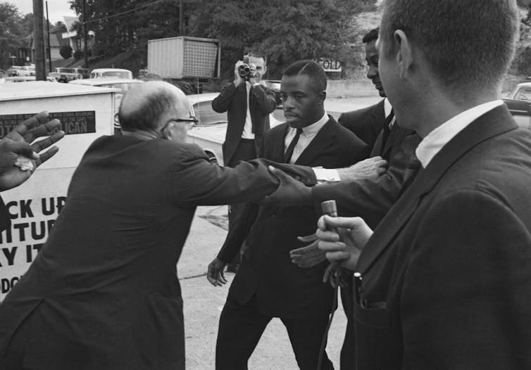 A bald white man pushes away a well-dressed black man from a restaurant.