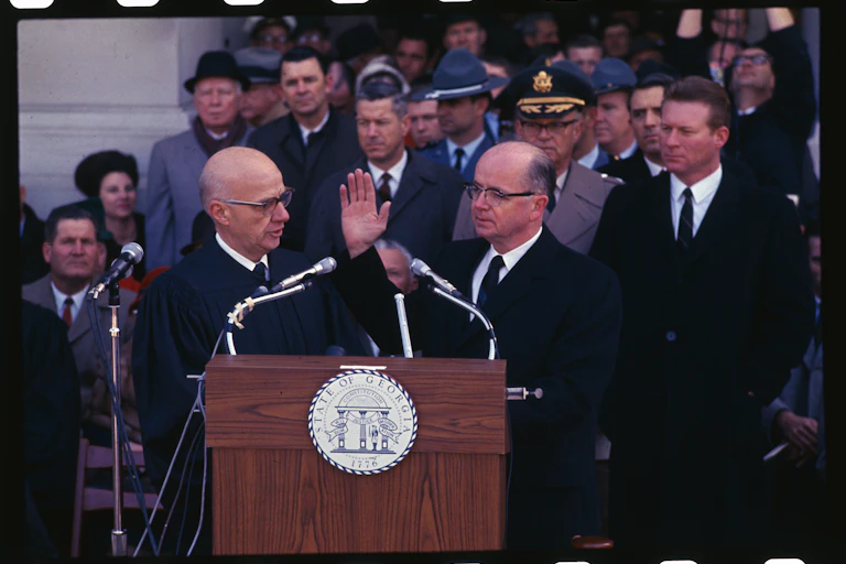 A white man raises his right hand as he is surrounded by other white men.