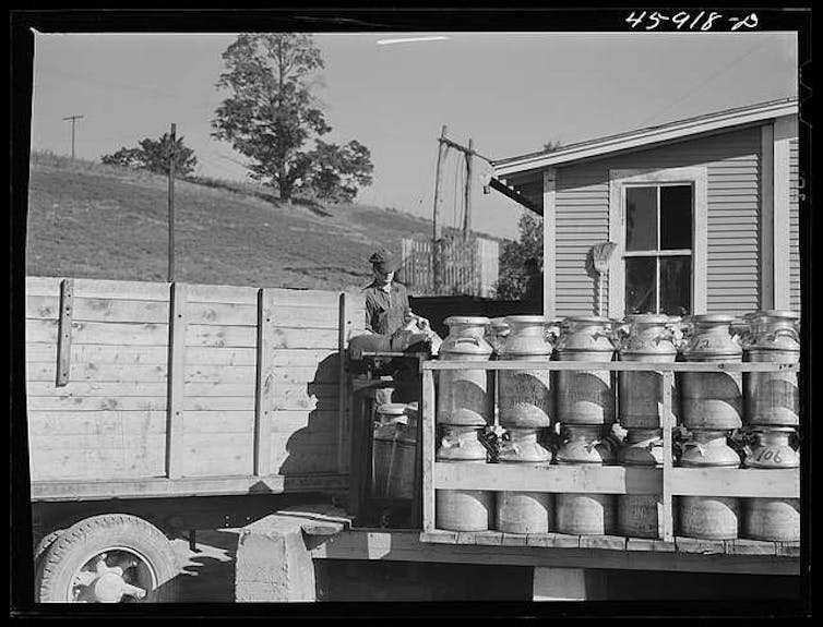 A man in work clothes stands on a truck bed loaded with stacked multi-gallon cans.