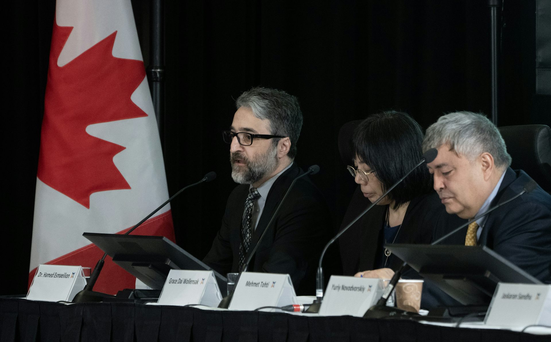 Two men with a woman sitting between them at a witness table with microphones and name plates in front of them and a Canadian flag to their right.
