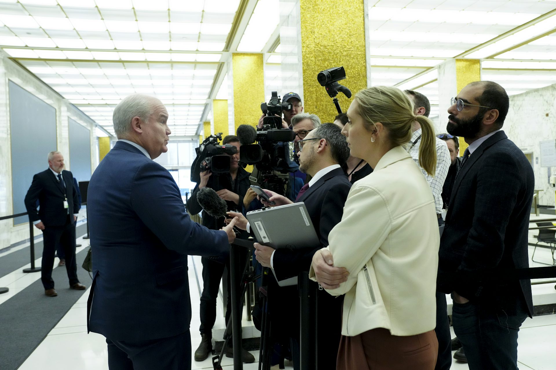 A bald man stands talking to a group of reporters in a brightly lit hallway.