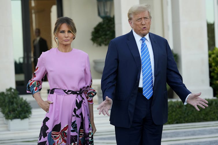 A rotund man with blond-grey hair in a blue suit and tie stands next to a frowning brunette woman in a pink dress.