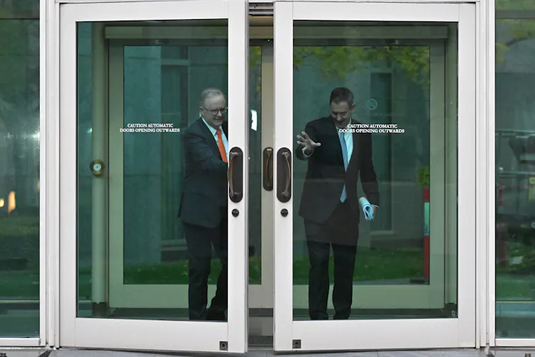 Two men in suits opening a large glass door in a long distance view.