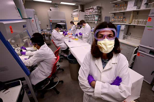woman with arms crossed in lab setting with many workers in PPE
