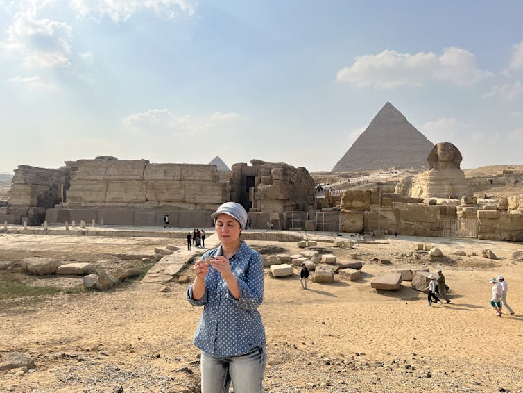 Photo of a woman standing on desert ground examining a piece of rock, with the Great Pyramid of Giza and the Sphinx in the background.