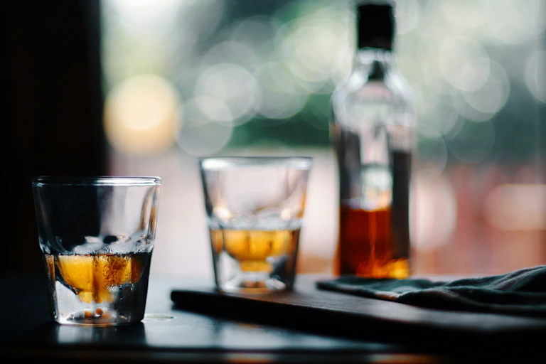 Two glasses half-filled with amber liquid sit next to a bottle of whiskey on a table.