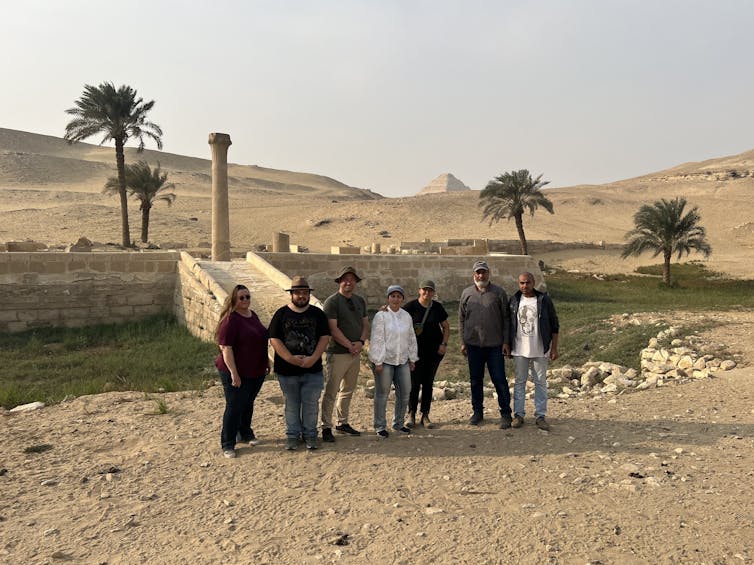A group of people standing in a desert in front of ancient stone steps leading up from a vegetated hollow to a further stone structure, with a pyramid in the distance.