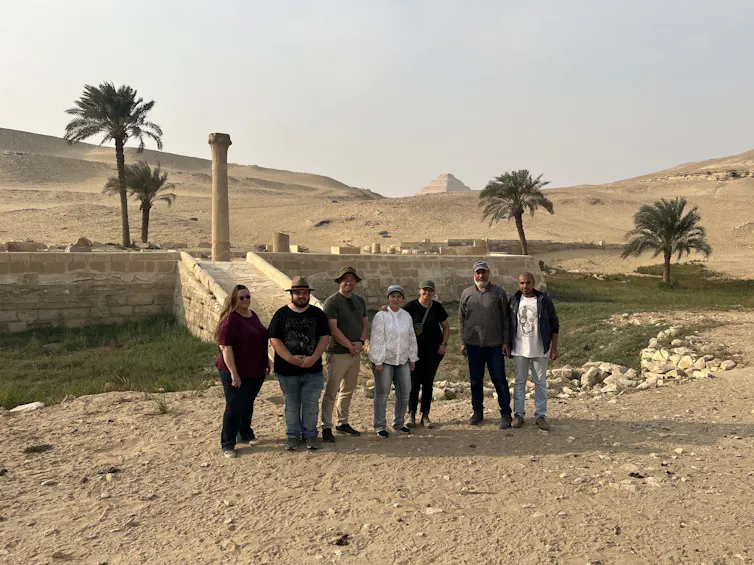 A group of people standing in a desert in front of ancient stone steps leading up from a vegetated hollow to a further stone structure, with a pyramid in the distance.