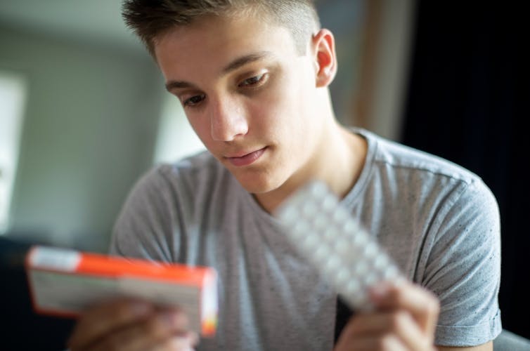 Teenage boy or young adult with blister pack of pills in one hand, holding and reading pill packet in the other