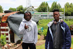 Two young men stand in front of a thriving vegetable garden, one gives the thumbs up!