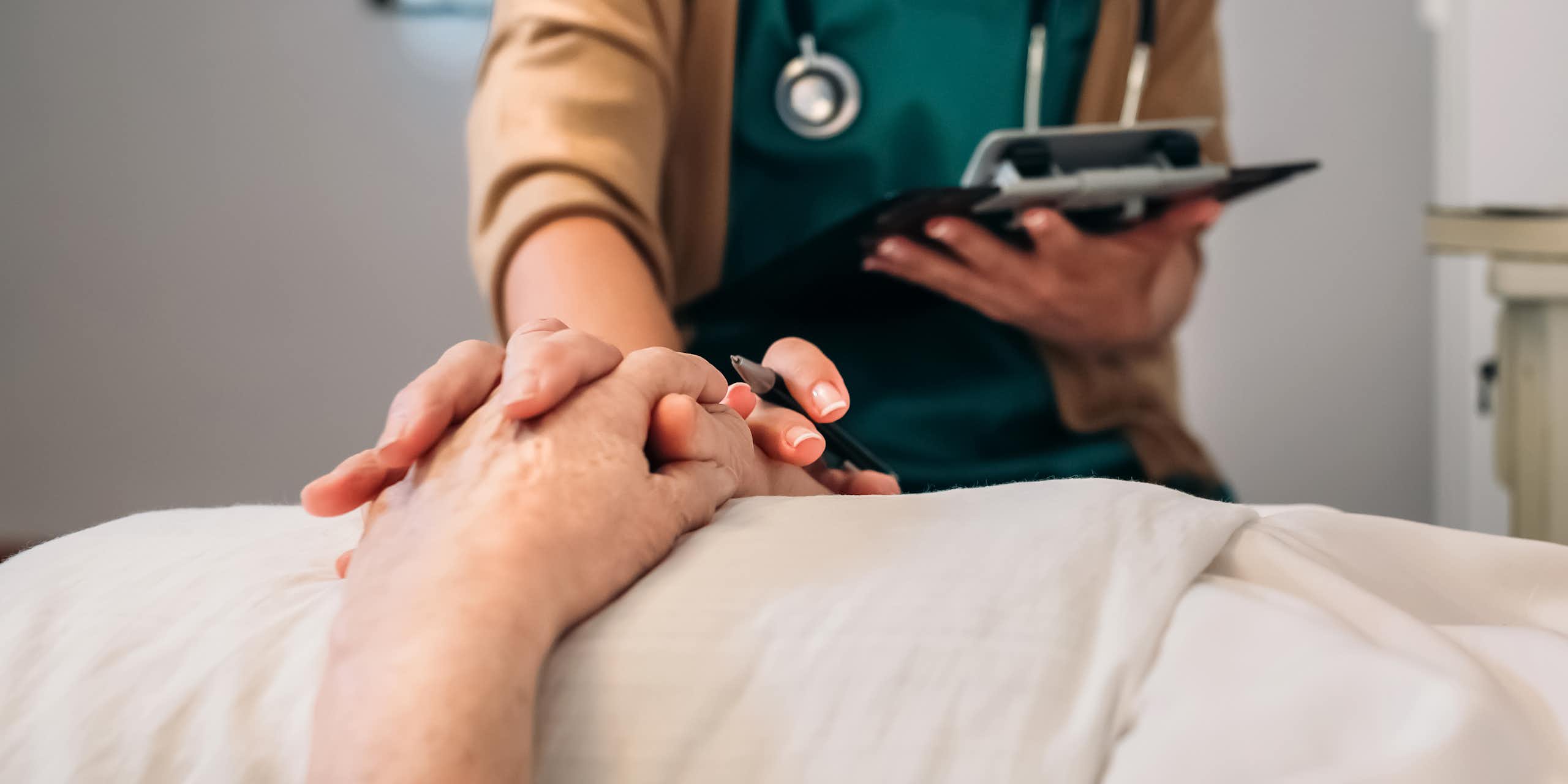 A doctor with a clipboard holds the hand of a patient on a bed.