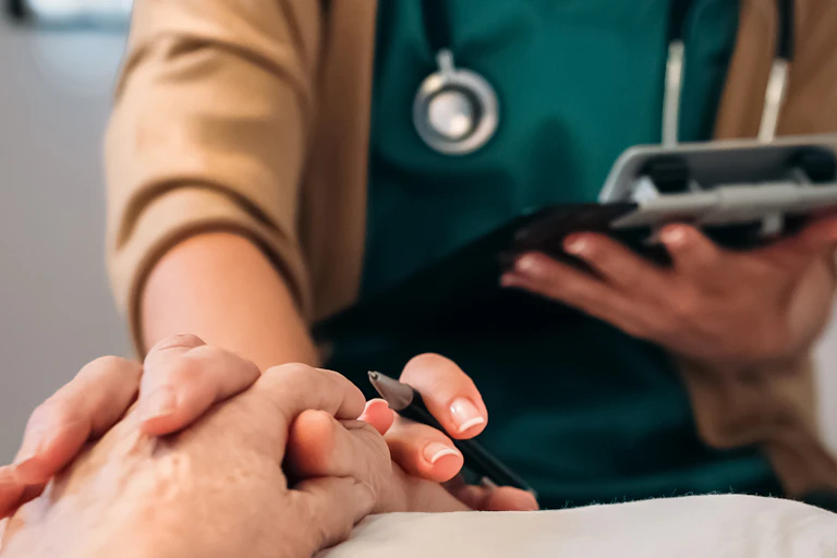 A doctor with a clipboard holds the hand of a patient on a bed.