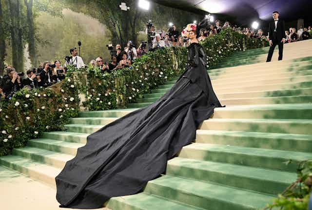 Zendaya wears a long black gown and a crown of flowers while standing on a sweeping staircase at the Met Gala.