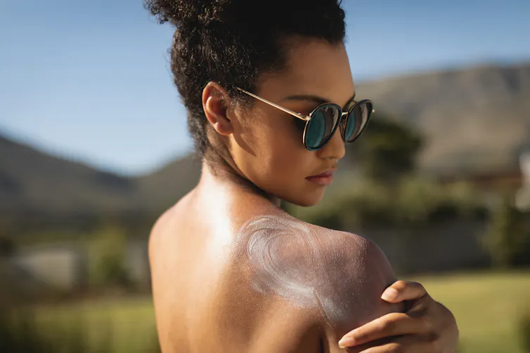 A woman applies sunscreen to her shoulder.