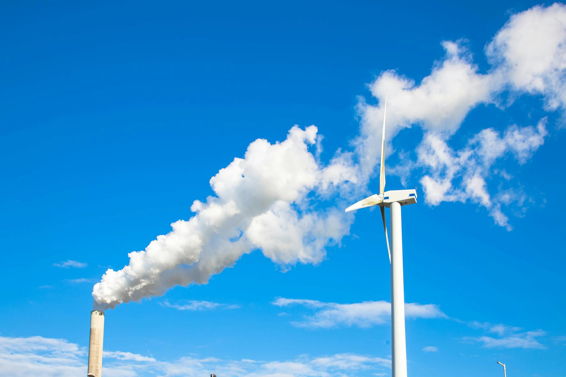 blue sky, white wind turbine on right, factory chimney on left with white smoke blowing across sky to right