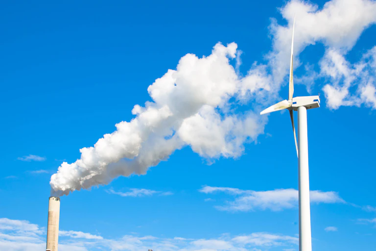 blue sky, white wind turbine on right, factory chimney on left with white smoke blowing across sky to right