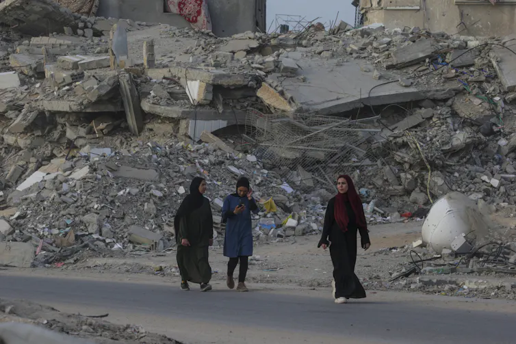 Three women walking down a road against the backdrop of a war-damaged city.