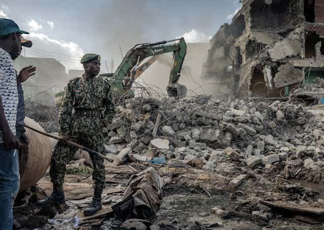 Man in military fatigue stands in front of rubble.