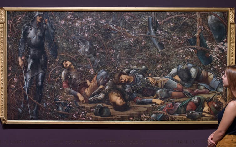 A large oil painting depicting fallen knights in armour is regarded by a woman in a museum.
