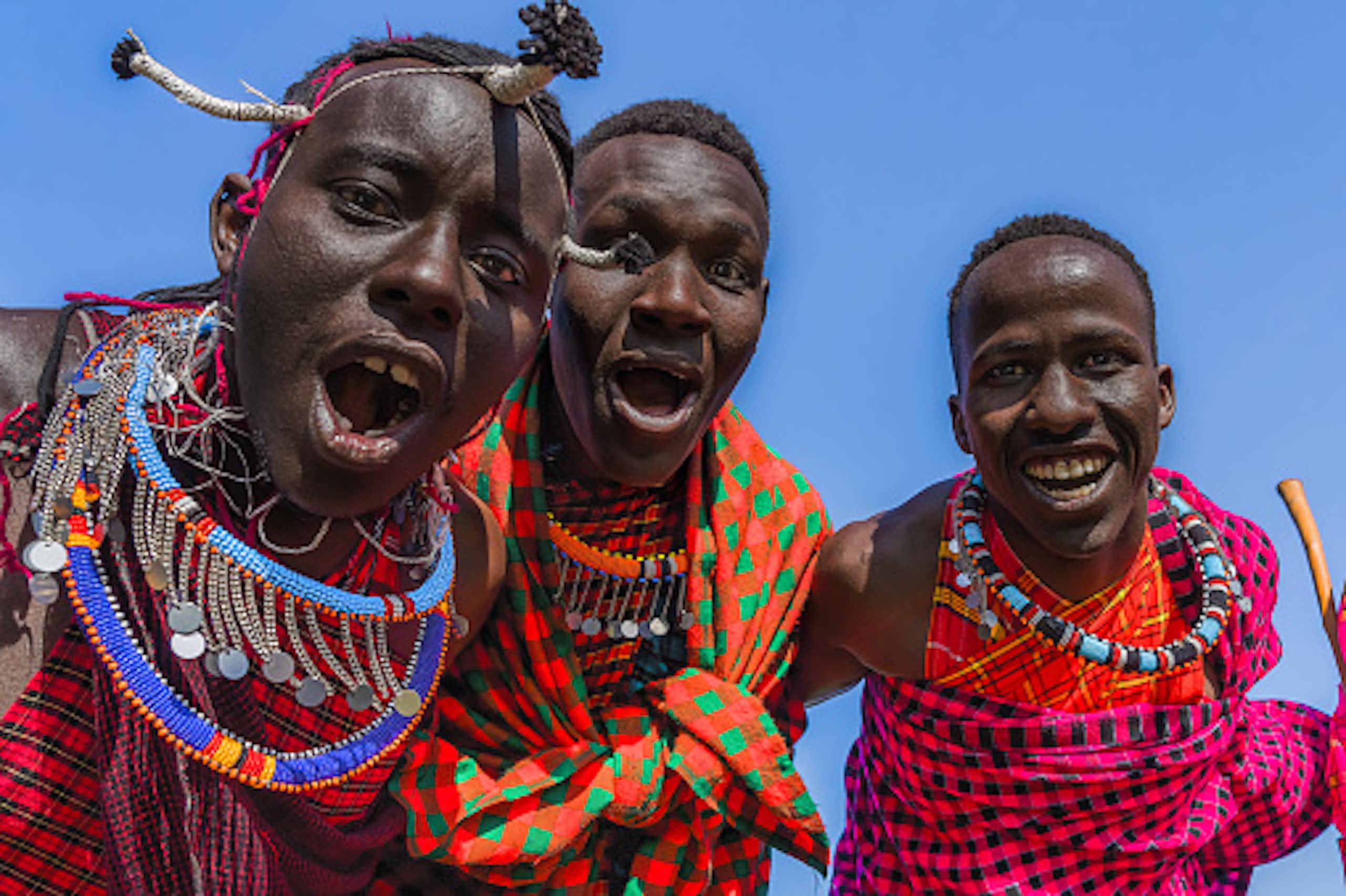Three young Maasai men in brightly coloured wraps and beads, leaning towards the camera with open mouths