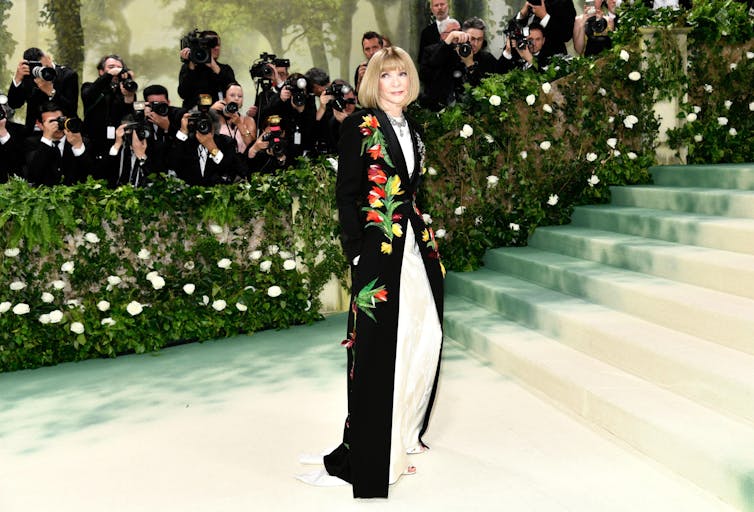 Anna Wintour stands at the bottom of a set of stairs in a long, black gown decorated with flowers.
