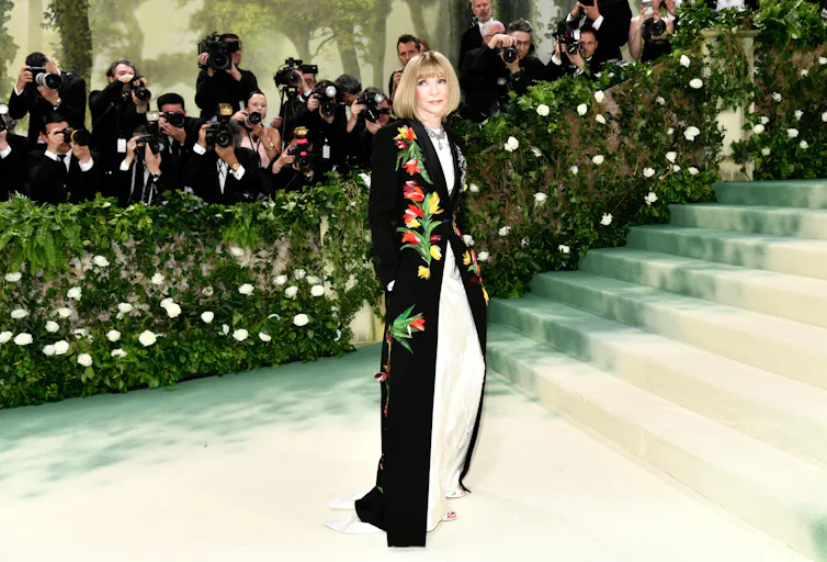 Anna Wintour stands at the bottom of a set of stairs in a long, black gown decorated with flowers.