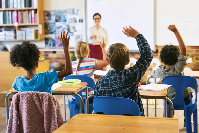 Des élèves de dos sont en classe et lèvent la main. L'enseignante est face à eux.
