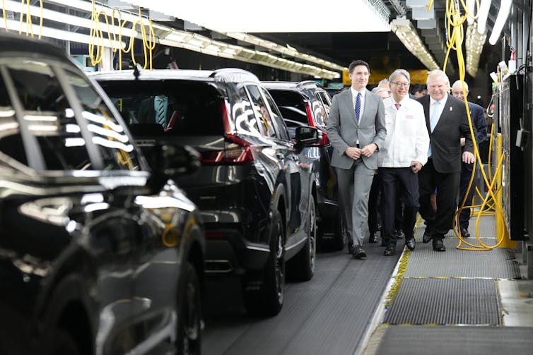 Three men walk past a column of cars in an automobile factory.