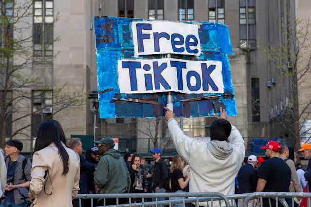 A person holds a handmade sign at the edge of a crowd outdoors in front of a large building