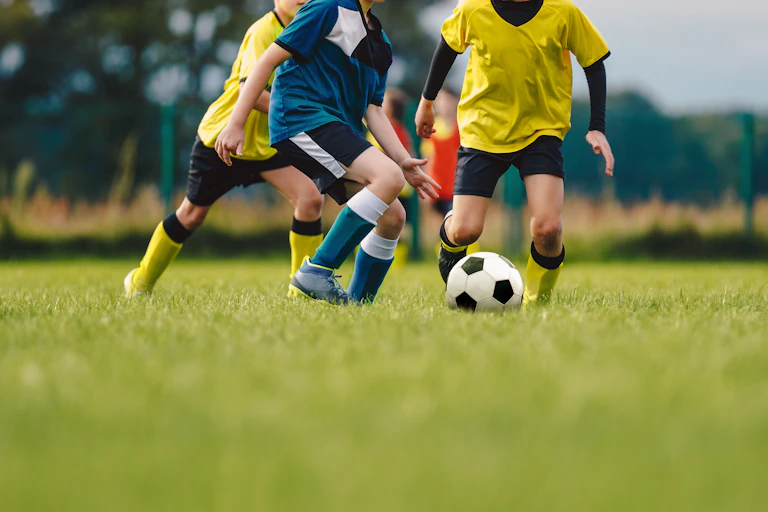 Children, seen from the neck down, kick a soccer ball on a grassy field while wearing soccer uniforms