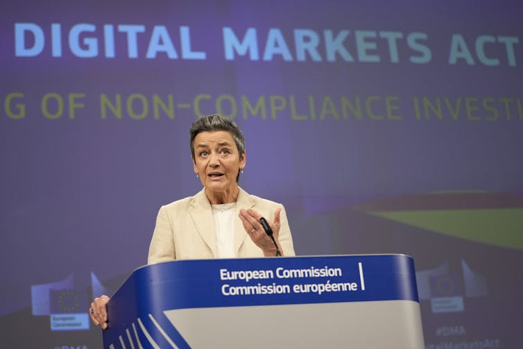 A woman stands in front of a large screen saying Digital Markets Act and speaks at a lectern