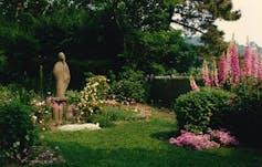 A statue of the Virgin Mary stands in the middle of a beautiful garden with colorful flowers, trees and plants.