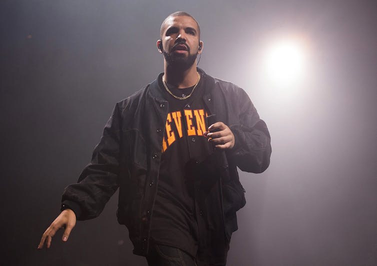 Drake wearing a black jacket and pants carries a mic while performing on a stage