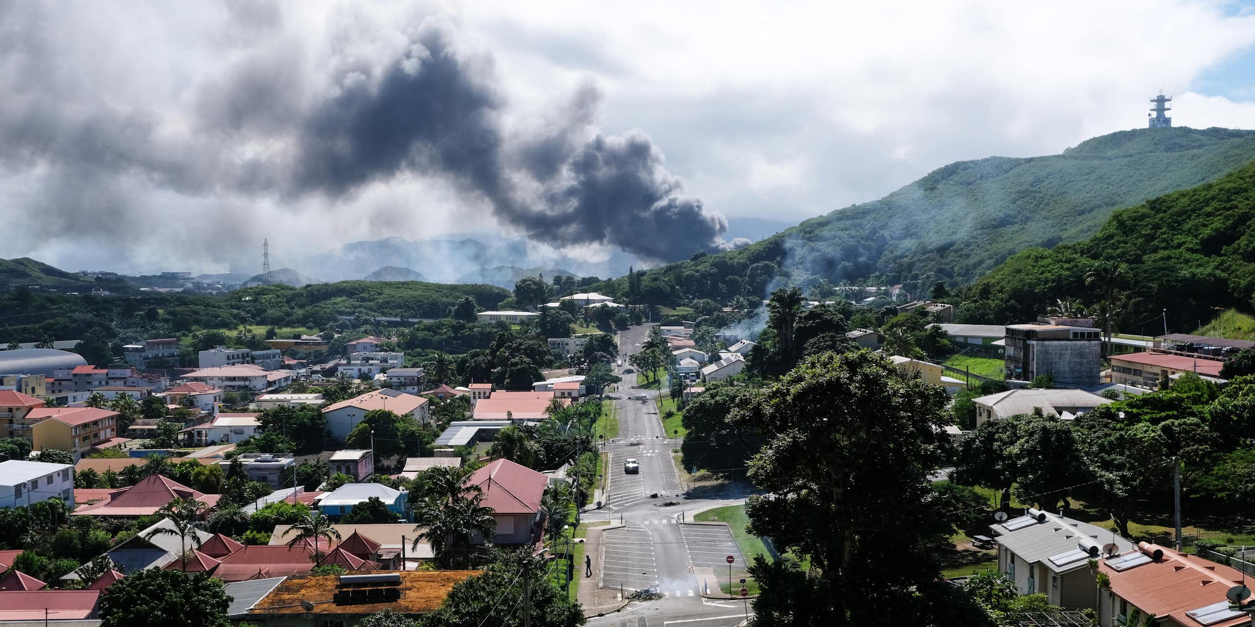 A drone view of a tropical township with plumes of smoke in the distance