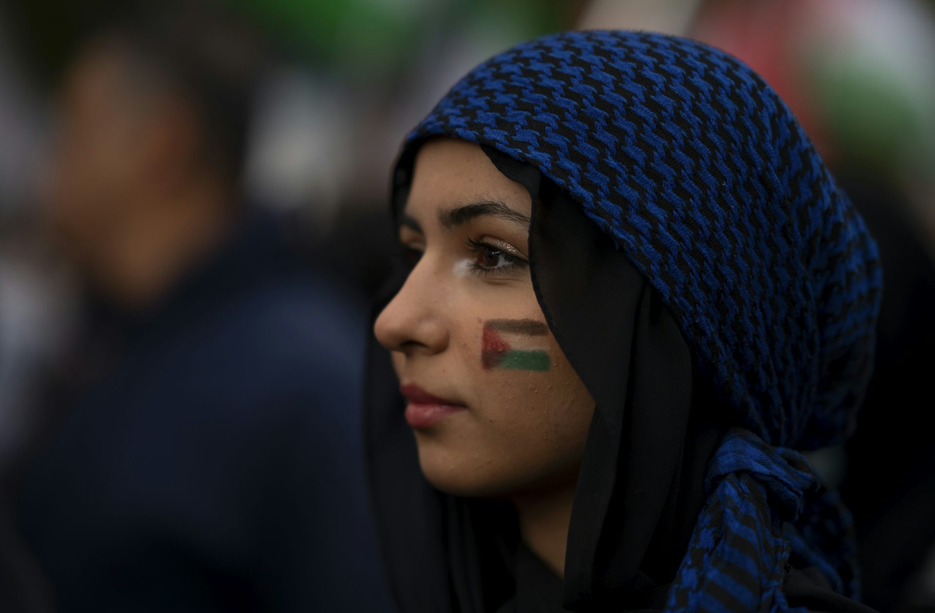 A woman wearing a blue keffiyeh around her head with a palestinian flag painted on her cheek