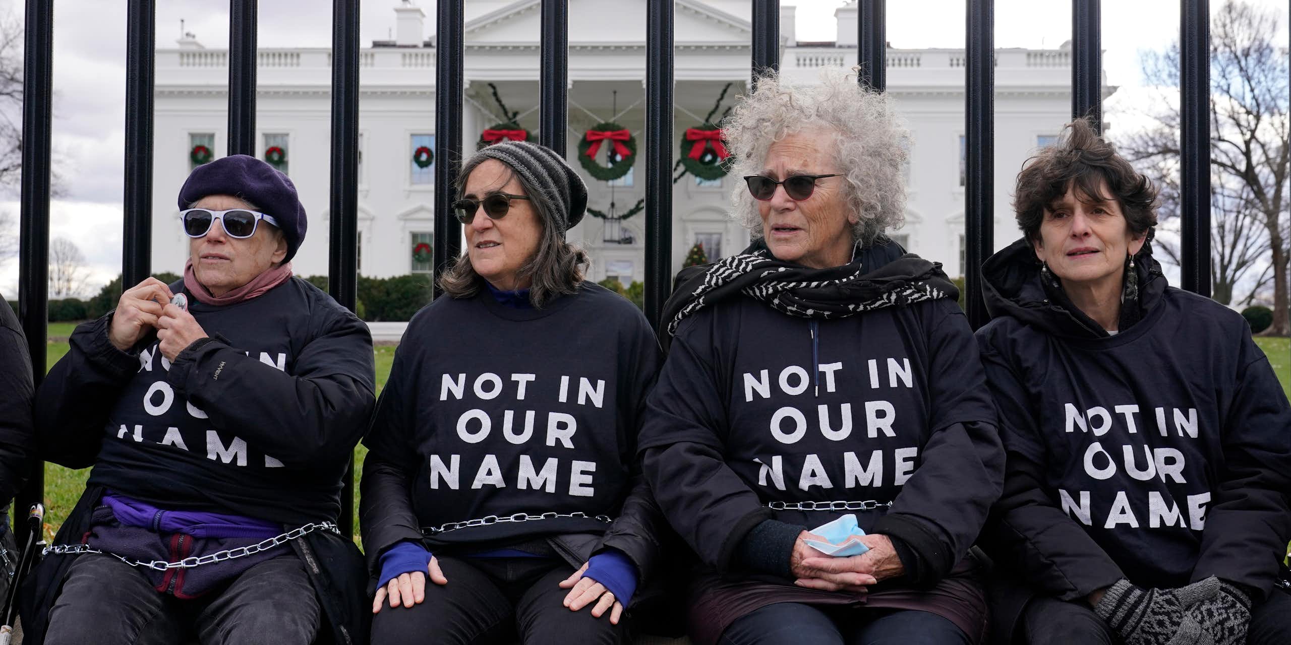 Four women in shirts that say 'Not in our name' sit on a fence with a large white mansion behind them.