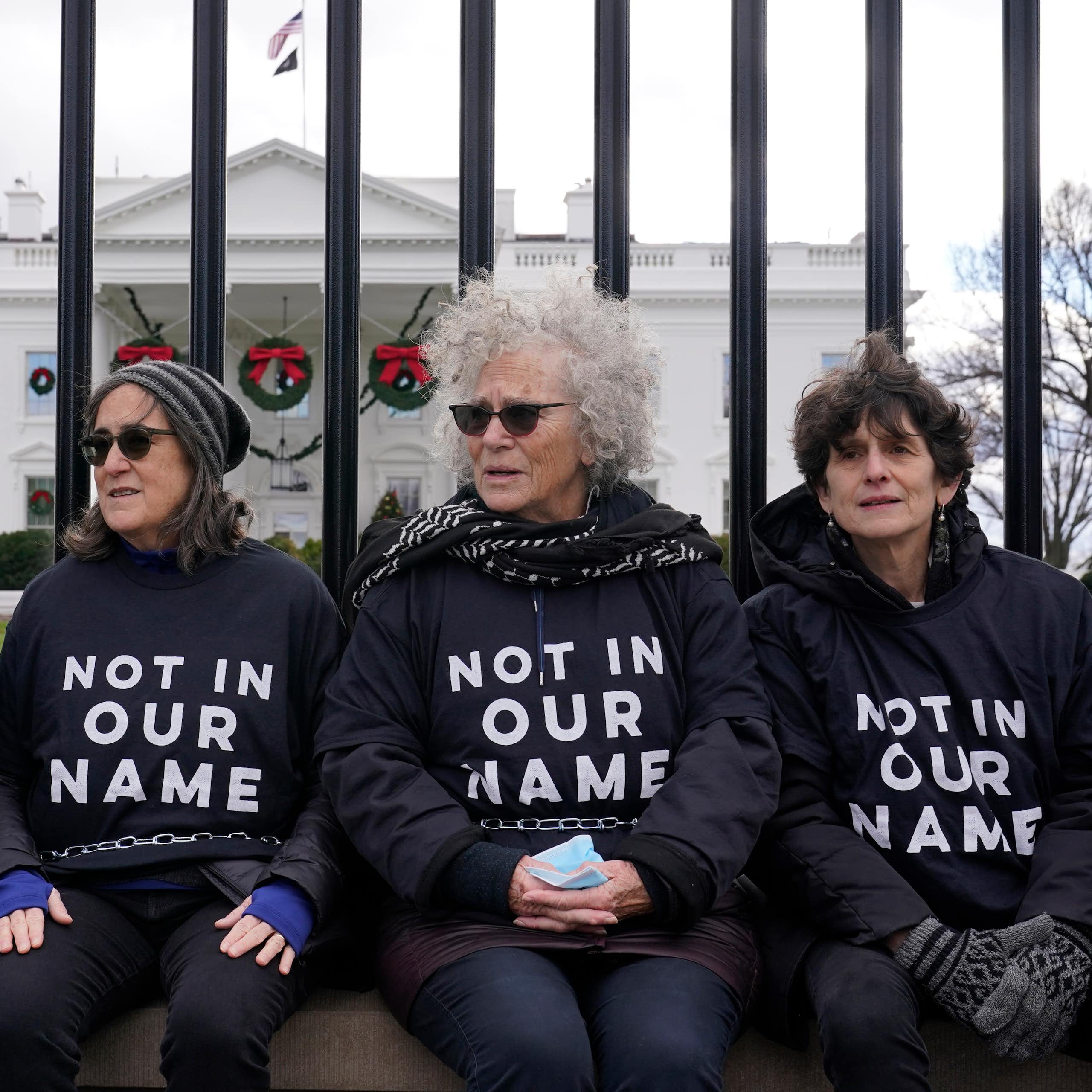 Four women in shirts that say 'Not in our name' sit on a fence with a large white mansion behind them.