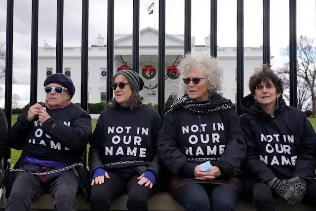 Four women in shirts that say 'Not in our name' sit on a fence with a large white mansion behind them.