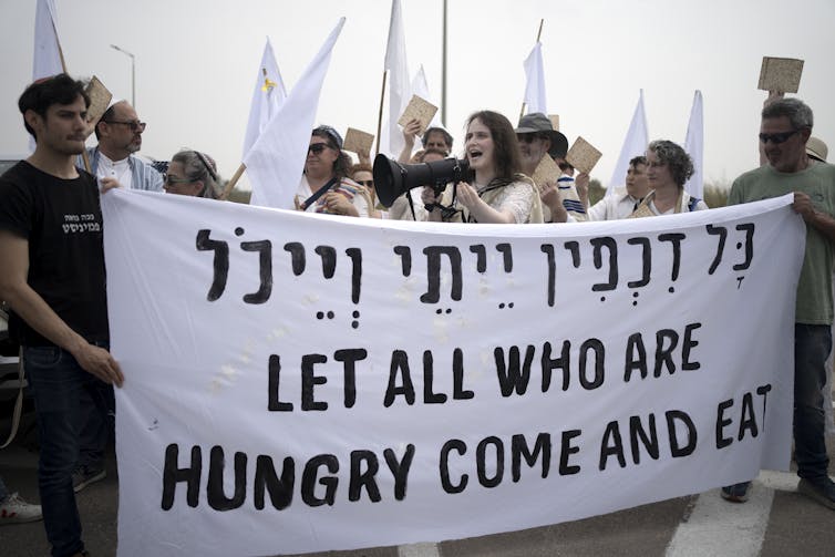 A woman speaks into a megaphone as other people hold a large white sign in Hebrew and English that reads: “All who are hungry, come and eat.”