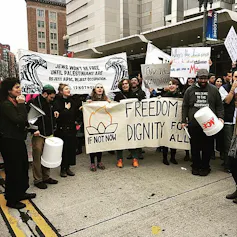 A group of protesters in coats hold white signs as they congregate on a sidewalk outside a city building.