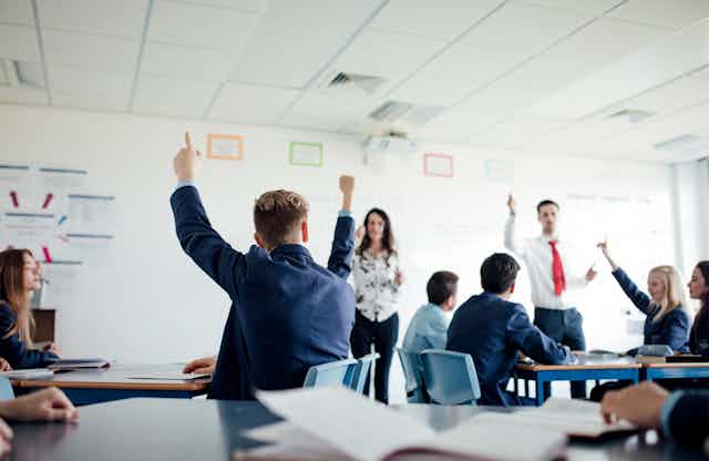 Students raise their hands in a classroom. Two teachers stand at the front of the students.