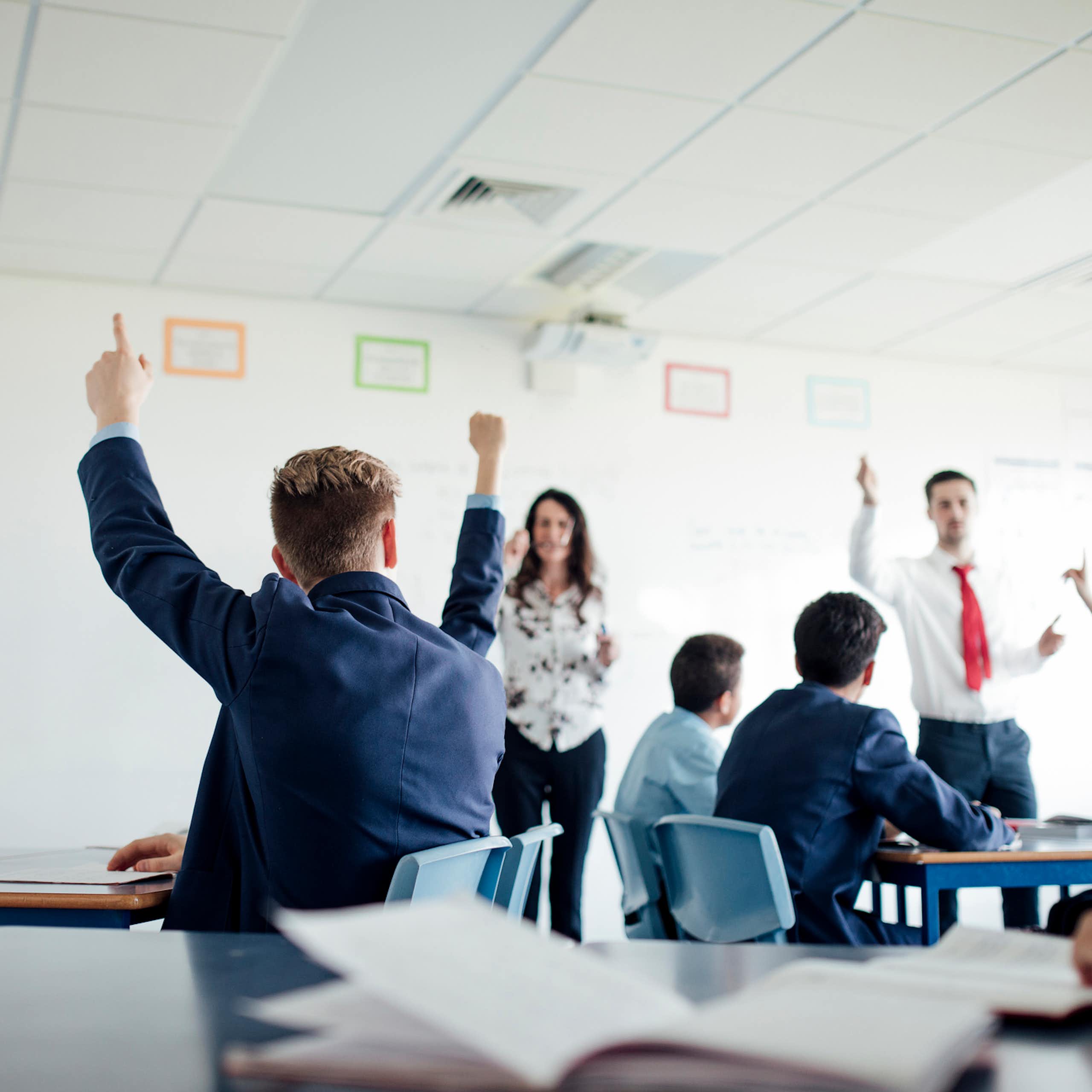 Students raise their hands in a classroom. Two teachers stand at the front of the students.