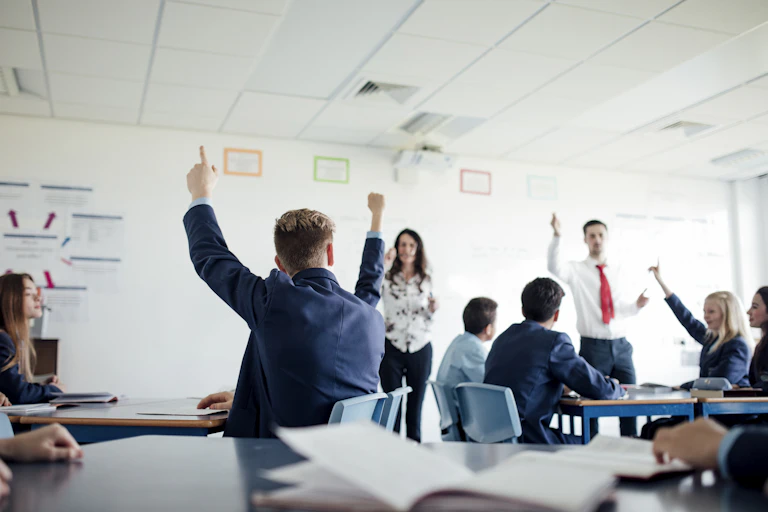 Students raise their hands in a classroom. Two teachers stand at the front of the students.